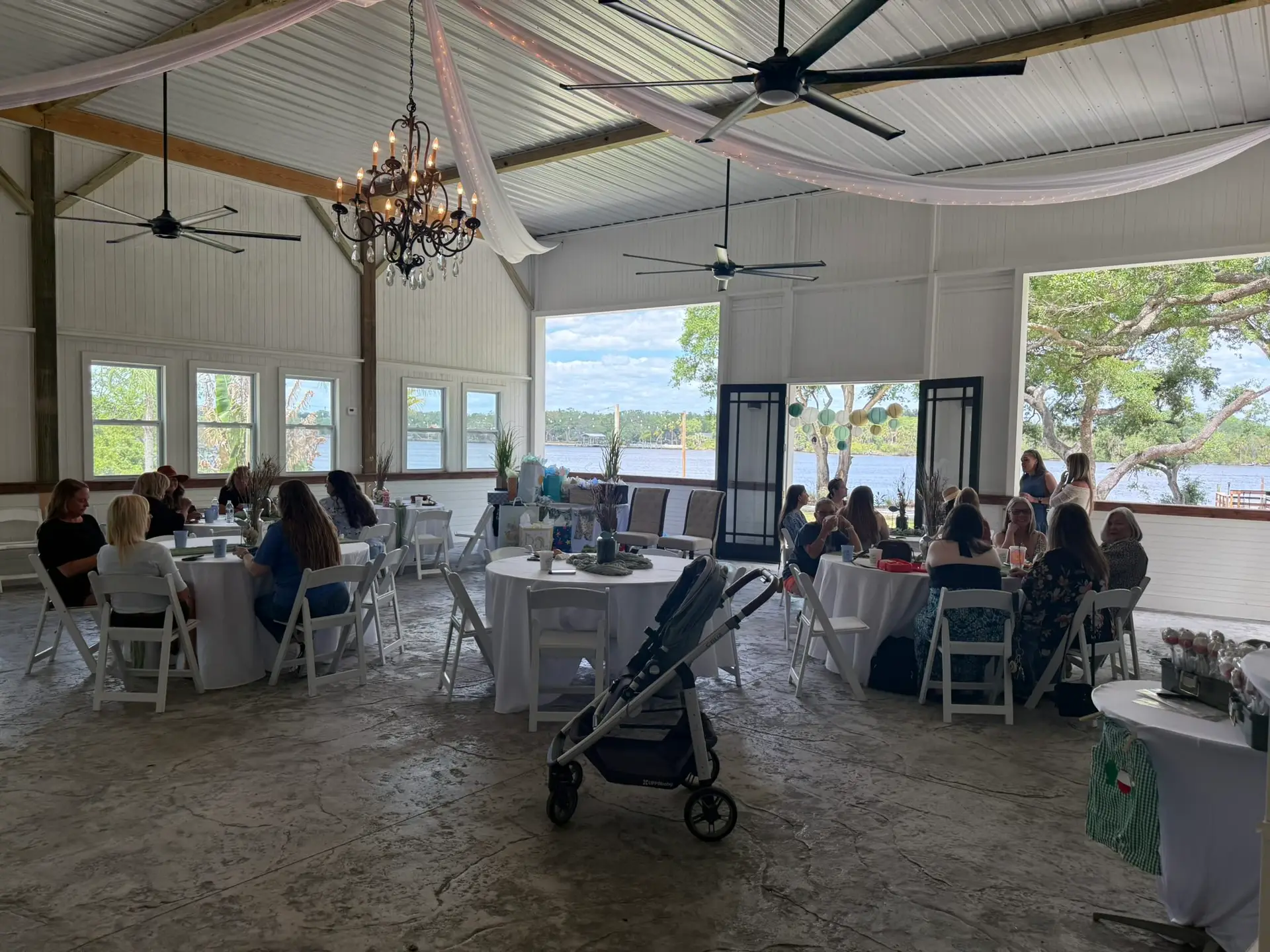 Bridal Shower at The Waterside Ranch - The Waterside Ranch Wedding reception in a bright barn by the water with round tables, white chairs, and a chandelier; open doors reveal a lake view.