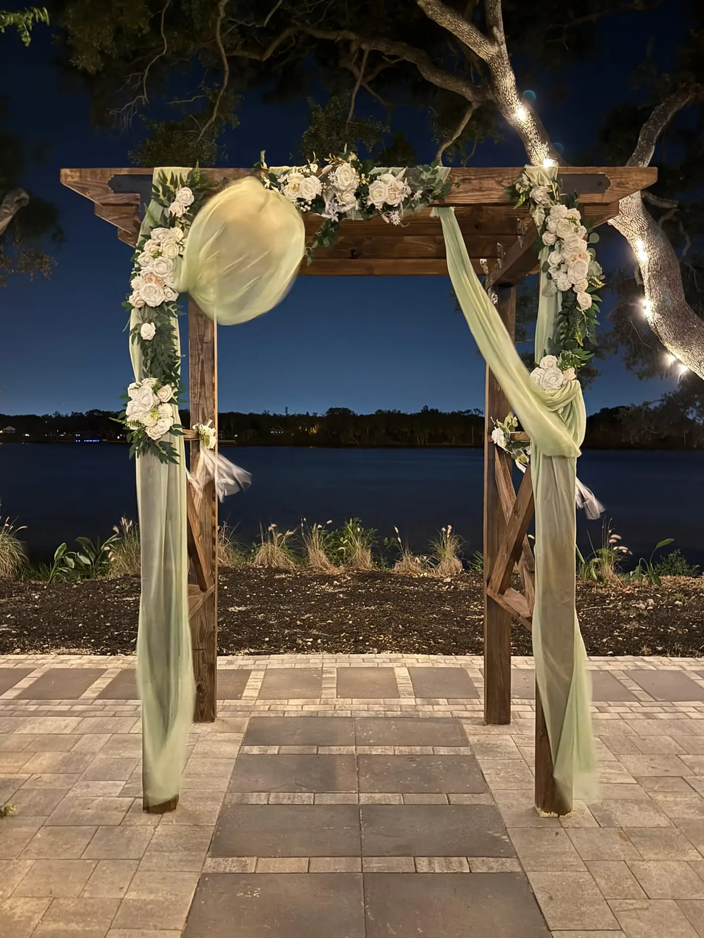 Ceremony Arbor - The Waterside Ranch - The Waterside Ranch Wooden wedding arch decorated with white roses and greenery, pale mint fabric draped along the sides, set by a body of water at night.