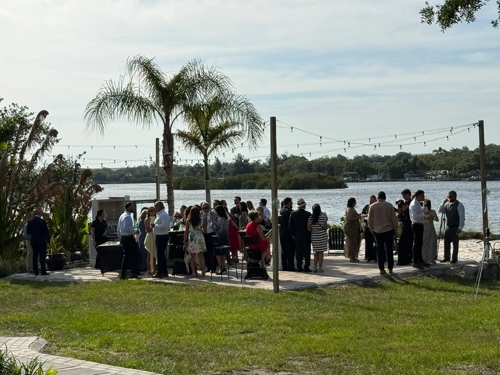 Courtyard Cocktail Hour - The Waterside Ranch - The Waterside Ranch Group of people in formal attire mingling outdoors by a lake, with palm trees and string lights overhead.