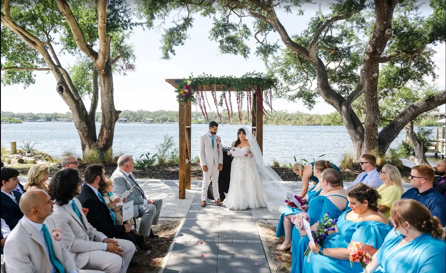 Bride and groom standing under a wooden floral arch by a lake as guests in light suits and turquoise dresses look on during an outdoor wedding.